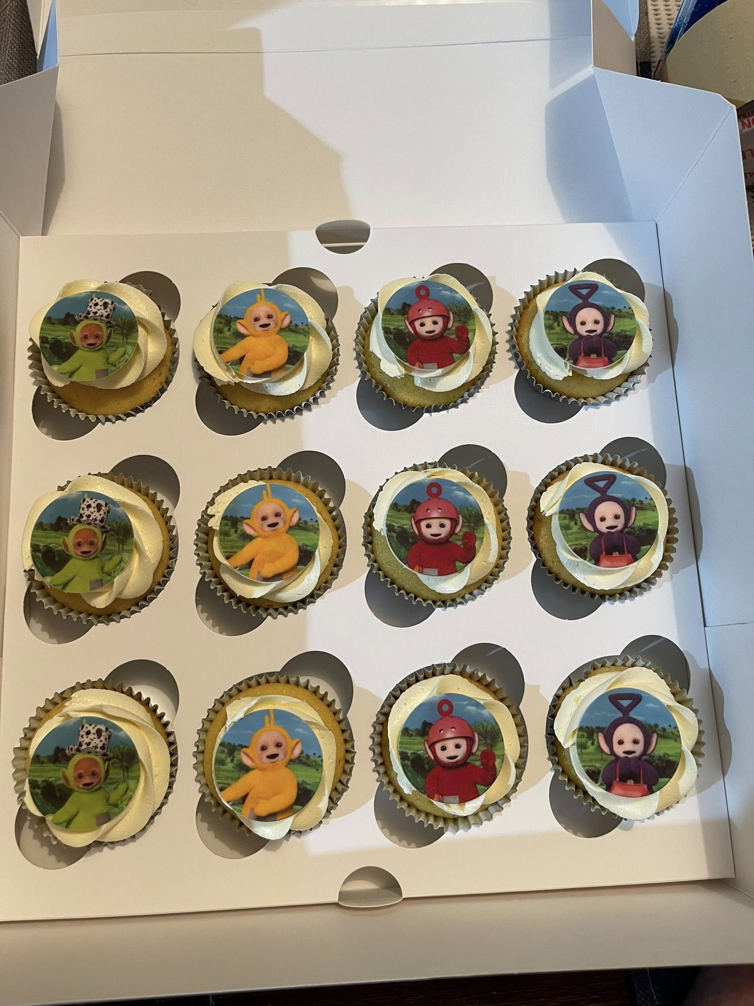 A dozen cupcakes with frosting topped with edible Teletubbies character images in a white box.