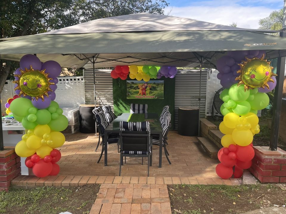 Teletubbies themed outdoor birthday party setup under a canopy with rainbow balloon pillars.