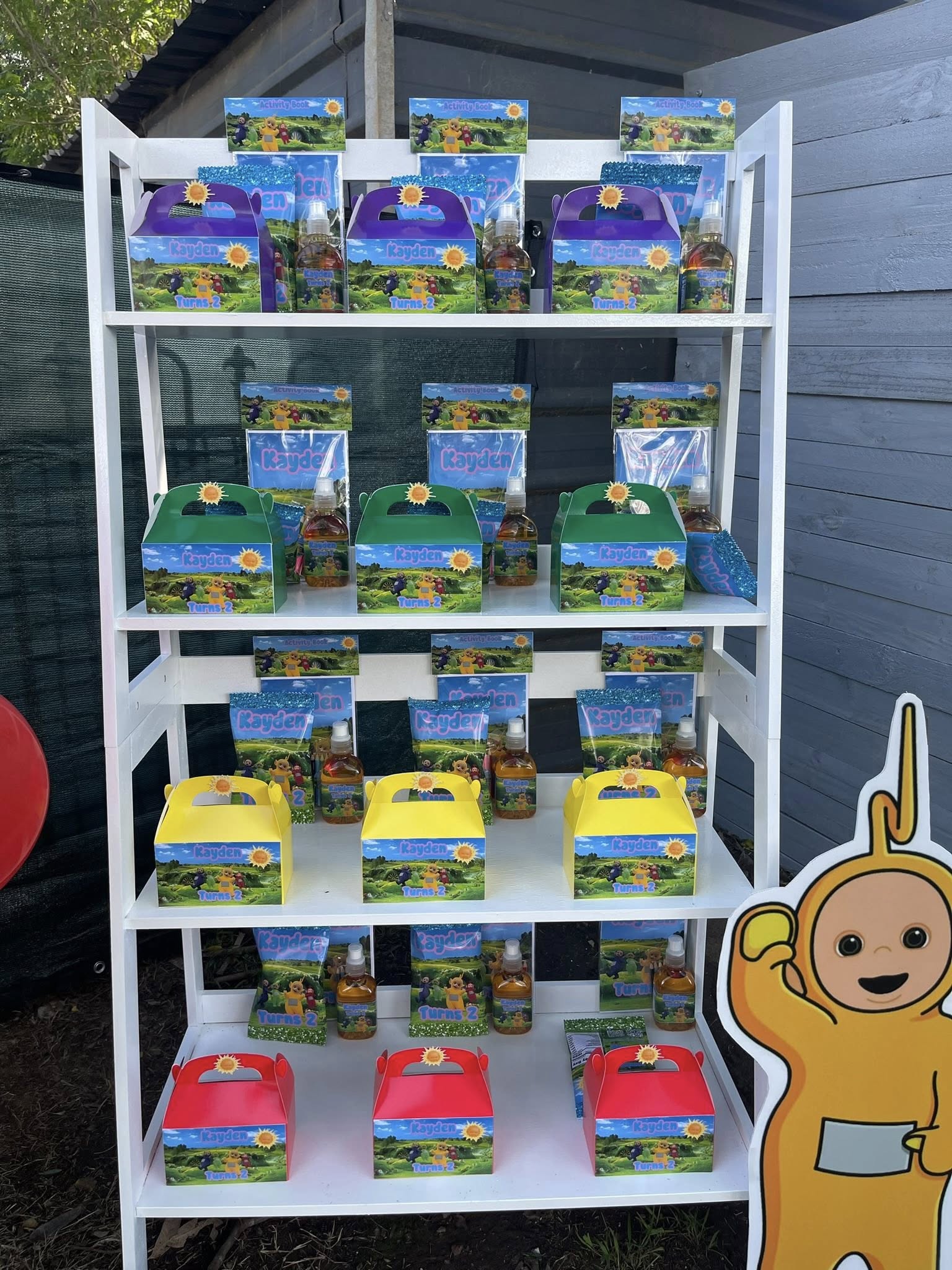 Shelves displaying personalized Kayden party favor boxes and customized Teletubbies activity books.