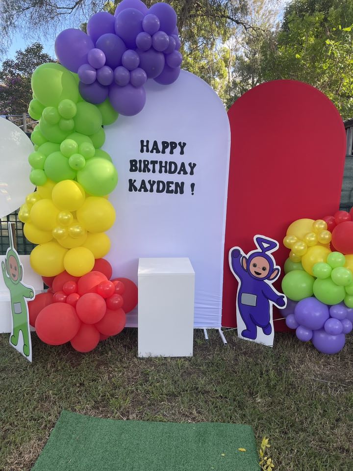 Teletubbies themed birthday setup with a colorful balloon arch and a Happy Birthday Kayden backdrop.