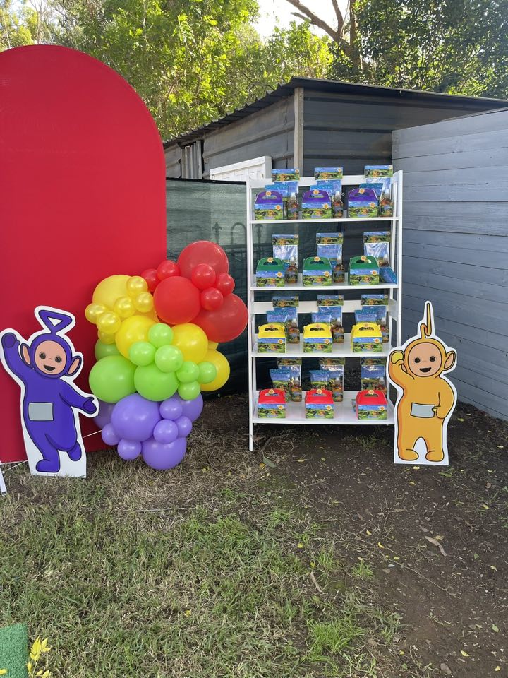 Teletubbies themed party display with balloon arch, character cutouts, and favors on a wooden shelf.