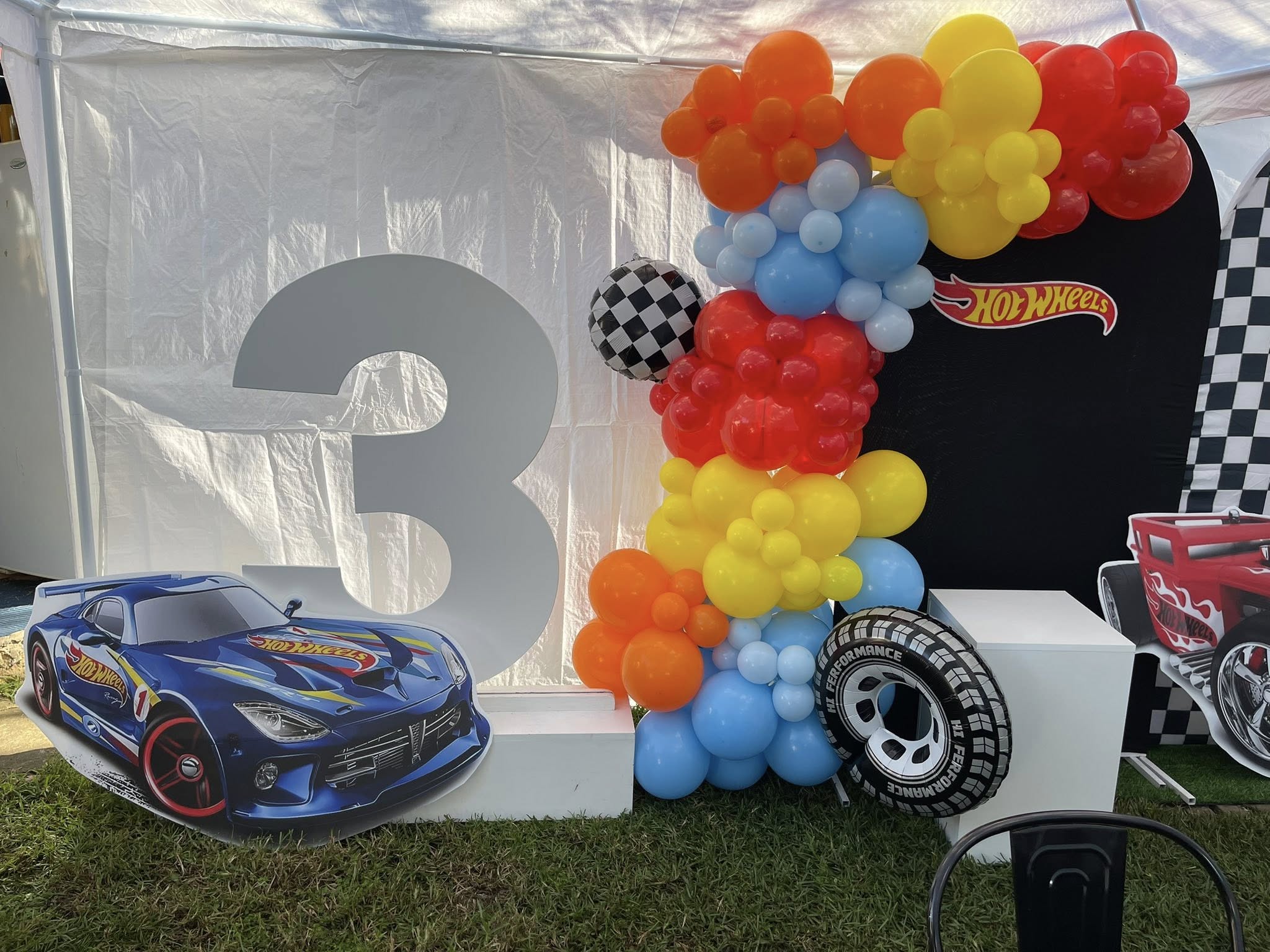 Hot Wheels themed third birthday party display with colorful balloon arch, number three prop, and racing car cutouts.