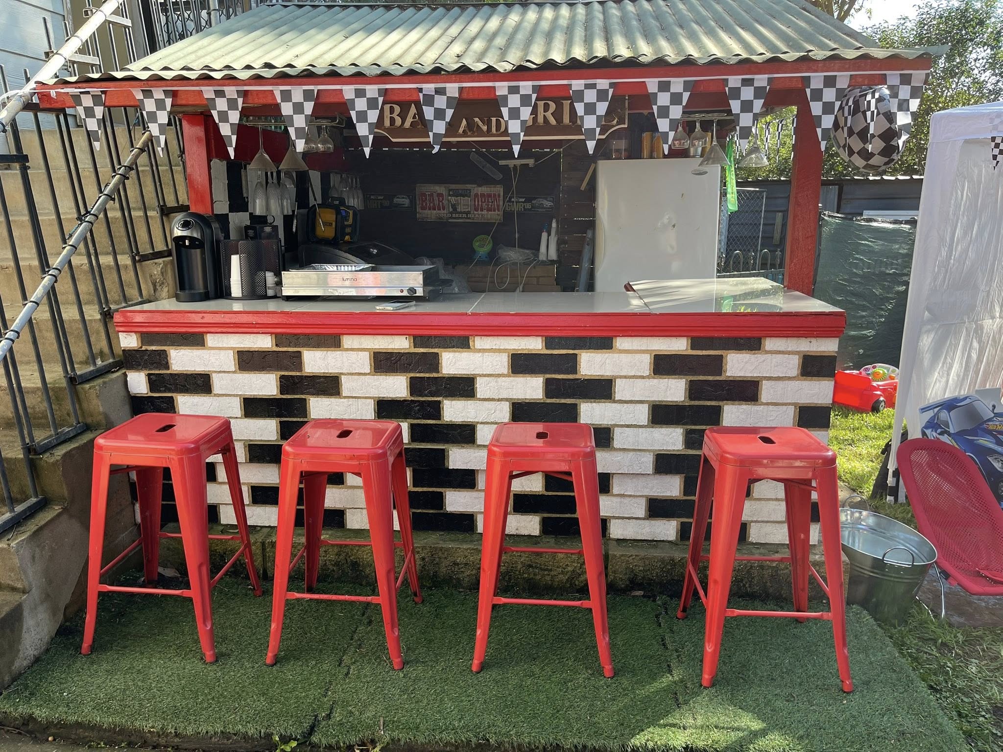 Outdoor backyard bar decorated with a racing checkerboard theme, featuring red bar stools and a brick-patterned counter.