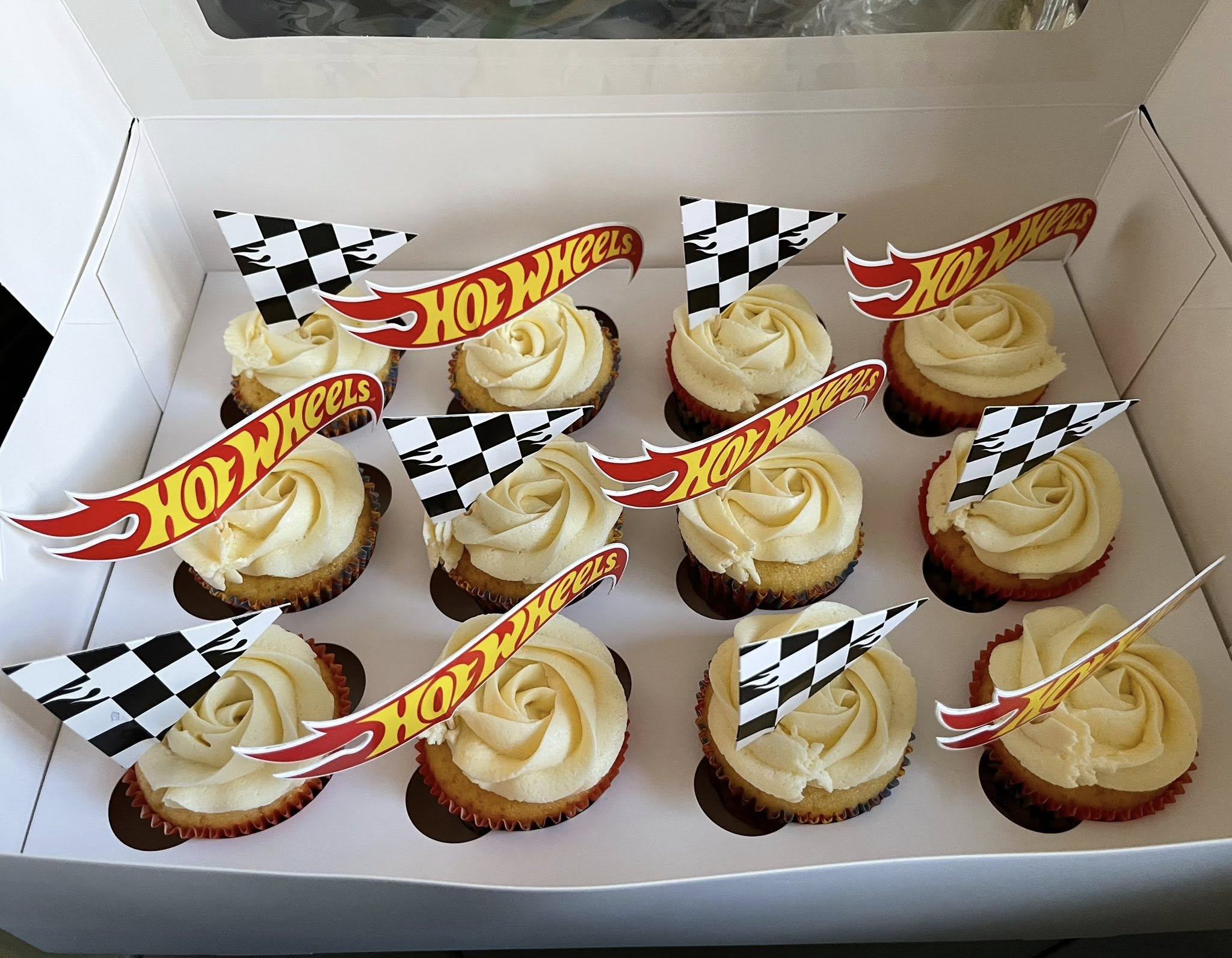 Dozen vanilla cupcakes with buttercream frosting, decorated with Hot Wheels checkered flags and logo toppers.
