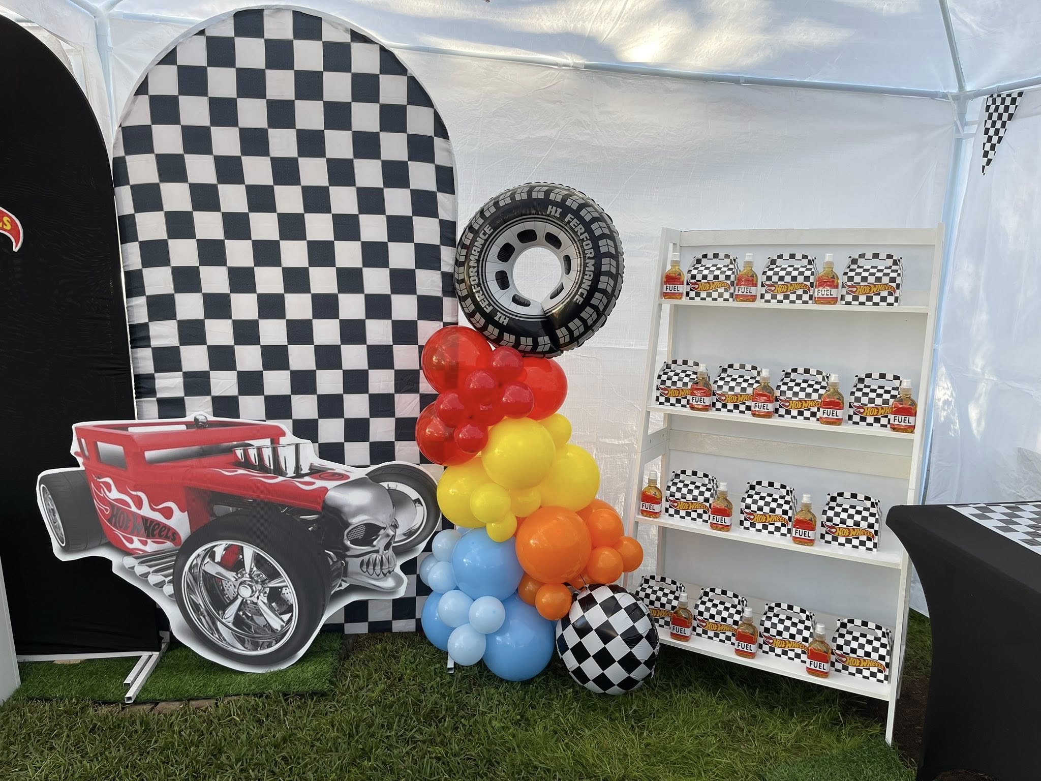 Hot Wheels themed party display with checkard backdrop, balloon arch, and party favors on a white shelf.