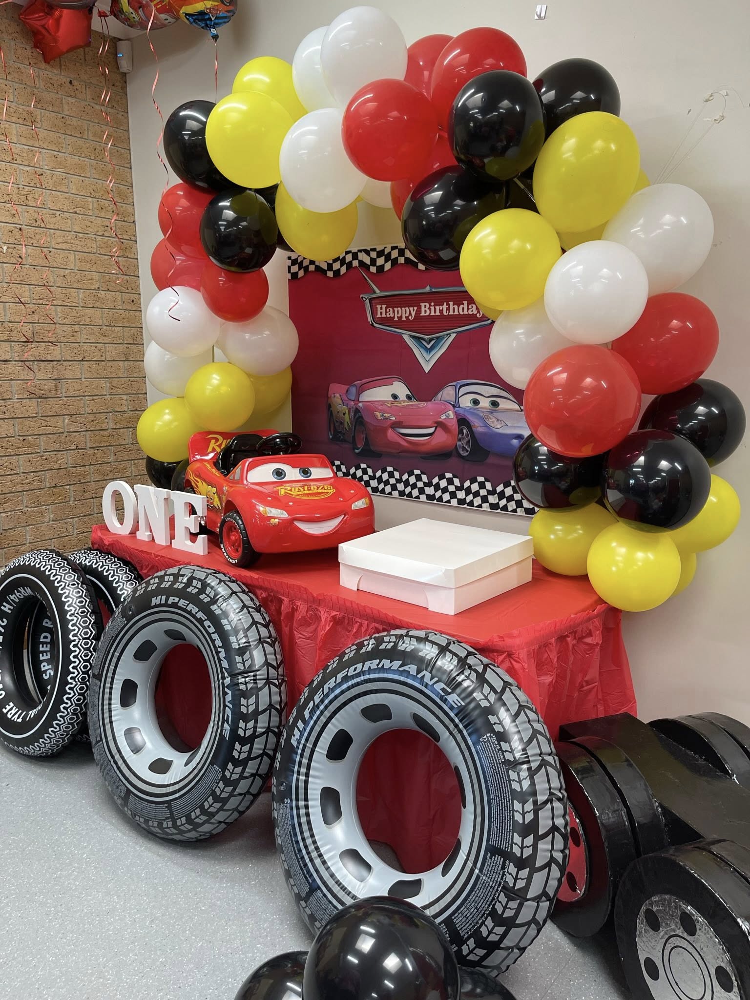 Cars themed first birthday party table decoration with balloons, Lightning McQueen car model, and tire props.