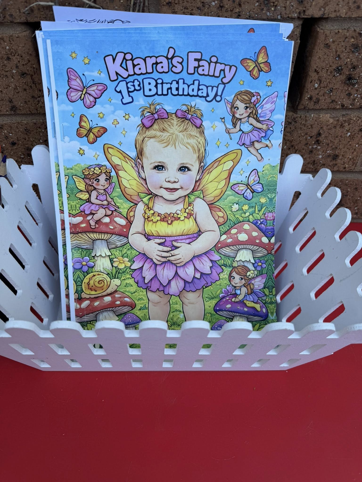 Personalized Kiara's Fairy 1st Birthday invitation cards in a white picket fence basket.