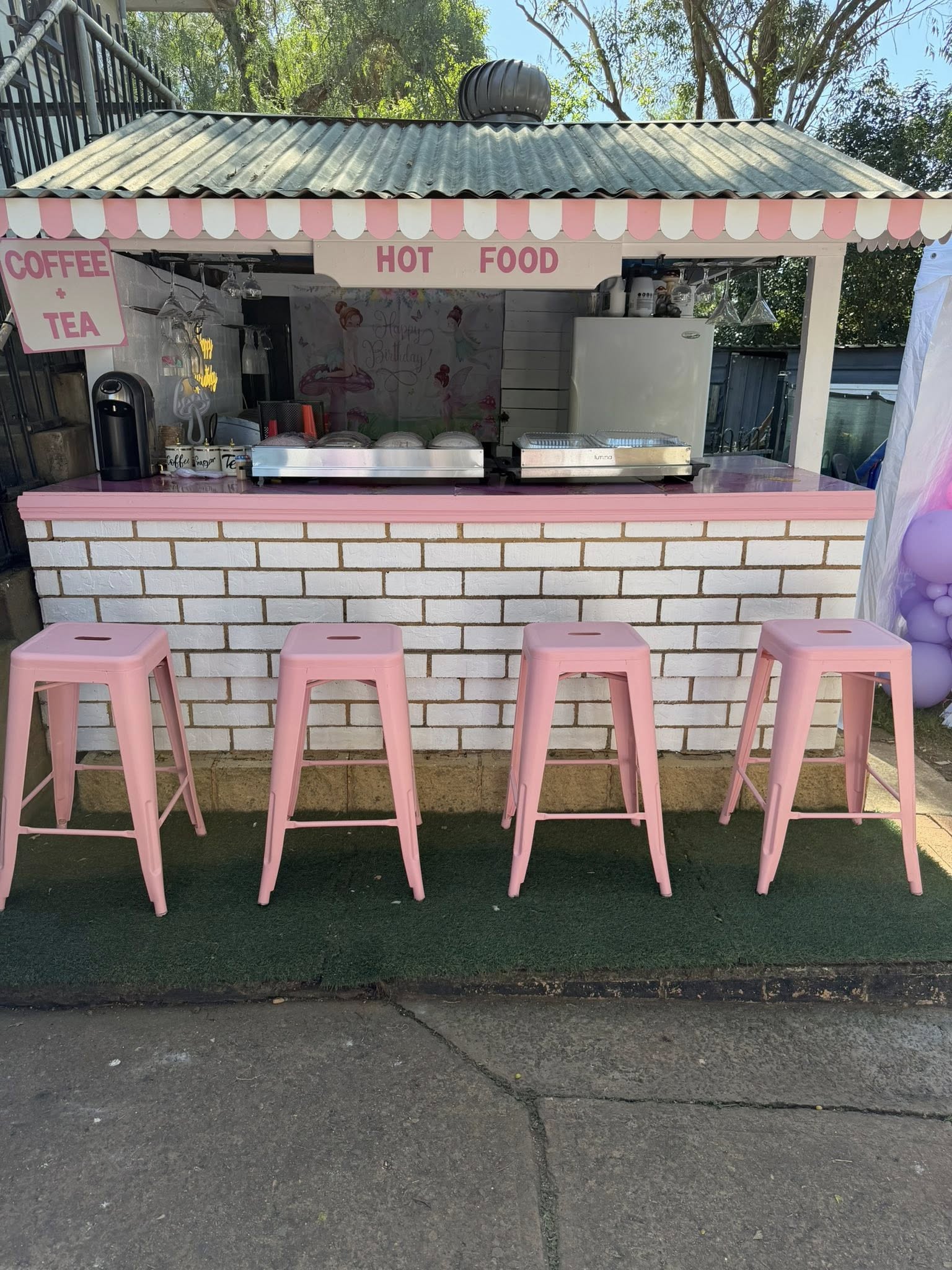 Outdoor catering food stall with a white brick base, pink trim, and four matching pink stools.