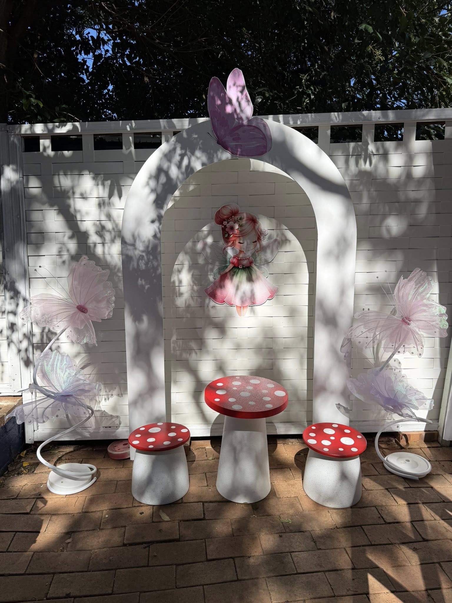 Fairy garden party backdrop with a white arch, mushroom-shaped table and stools, and sheer butterfly decorations.