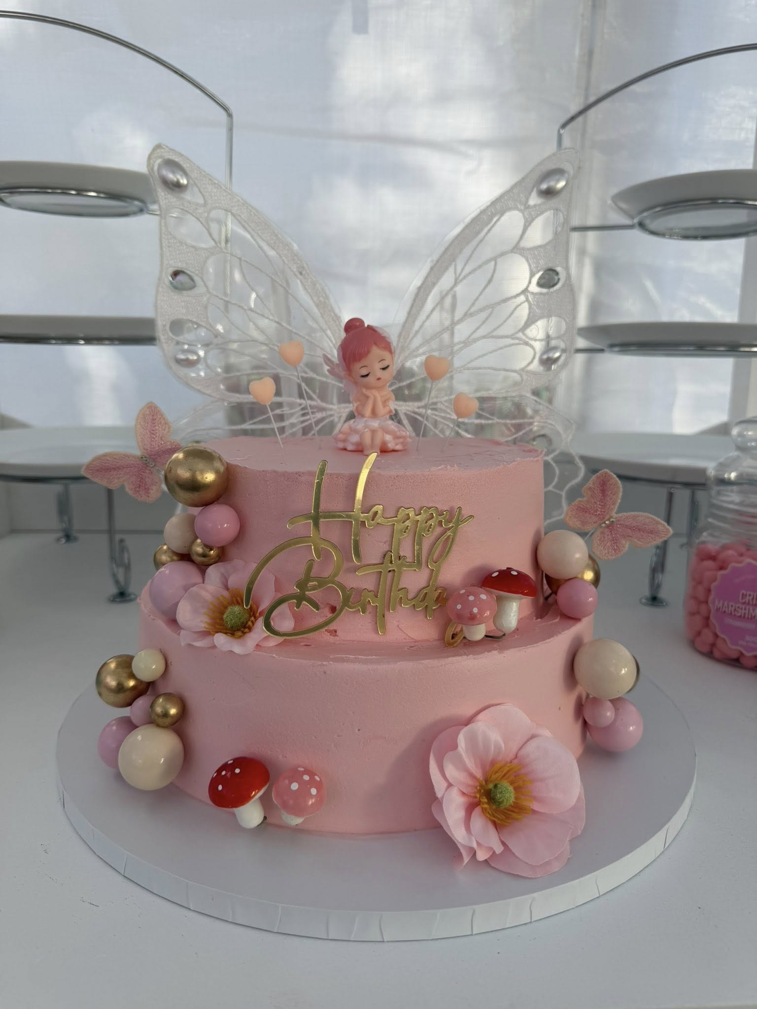 Two-tier pink birthday cake decorated with a fairy figurine, large wings, flowers, and mushroom accents.
