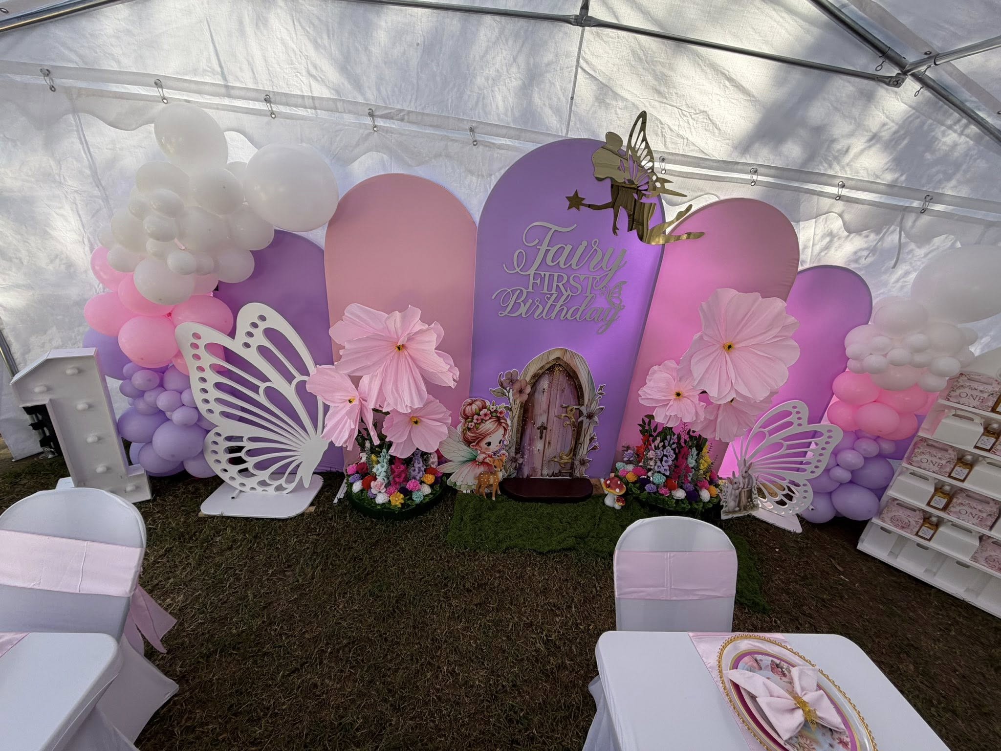 Fairy-themed first birthday party backdrop with pink and purple panels, oversized paper flowers, and balloon garlands.