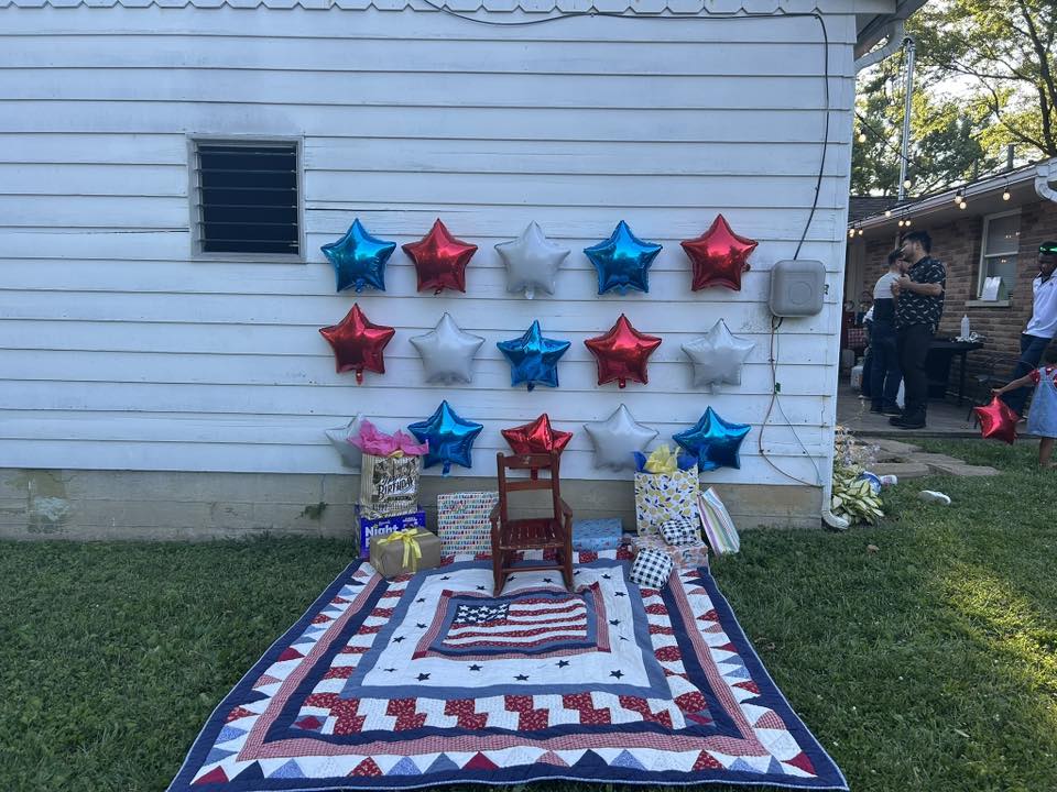 Patriotic outdoor birthday party setup with red, white, and blue star balloons, gifts, and a USA flag quilt.