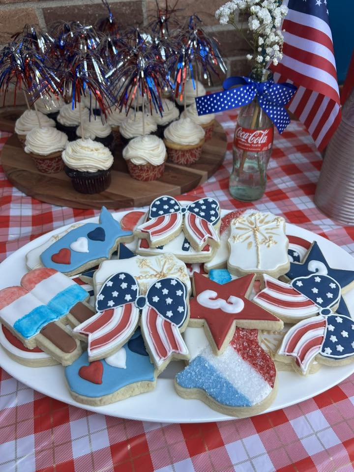 Platter of Fourth of July themed iced cookies shaped like stars, popsicles, and bows with patriotic cupcakes in the background.