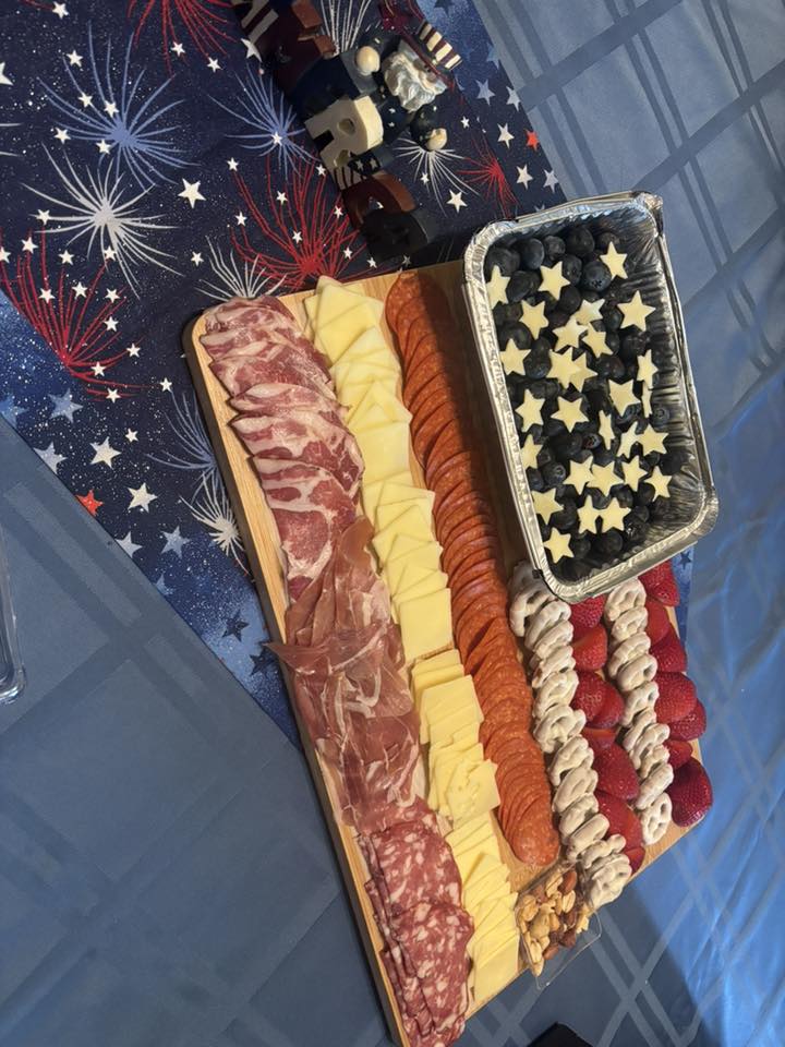 Patriotic charcuterie board featuring meats, cheeses, star-shaped fruit, and nuts on a firework tablecloth.