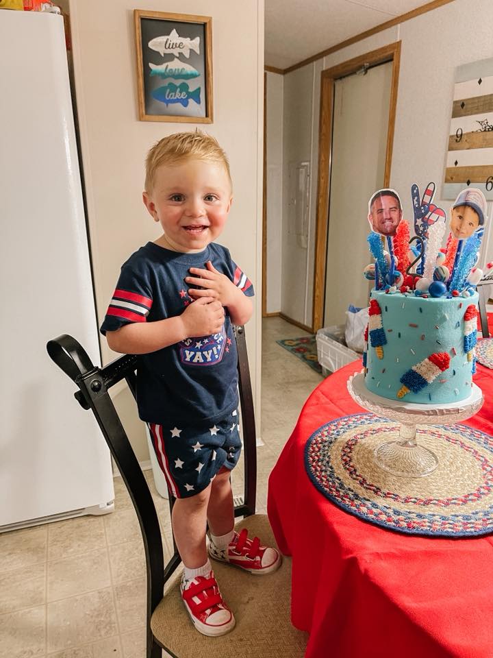 A smiling blonde toddler wearing a red, white, and blue patriotic outfit standing next to a birthday cake.