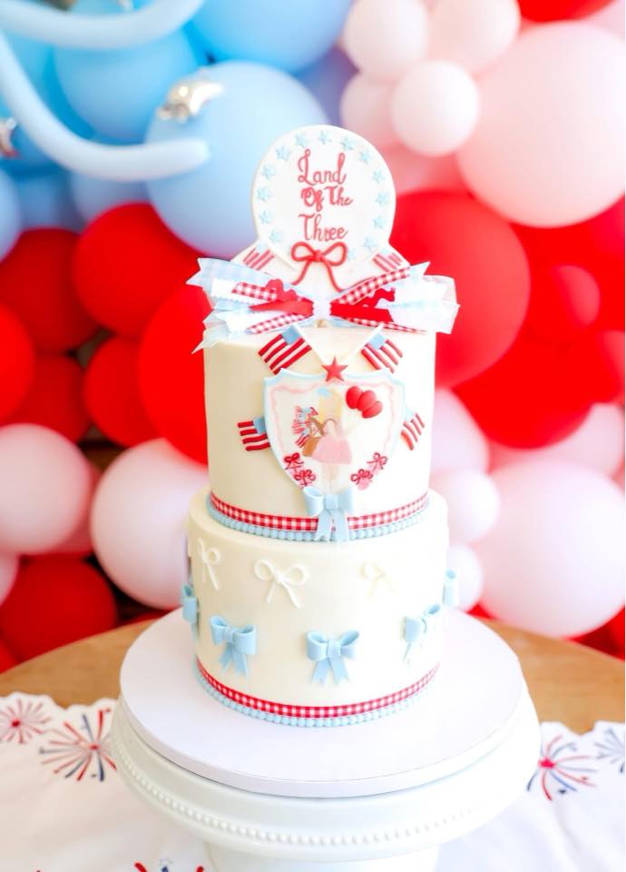 Two-tier tiered birthday cake themed Land of the Three with red and blue accents, bows, and patriotic decorations.