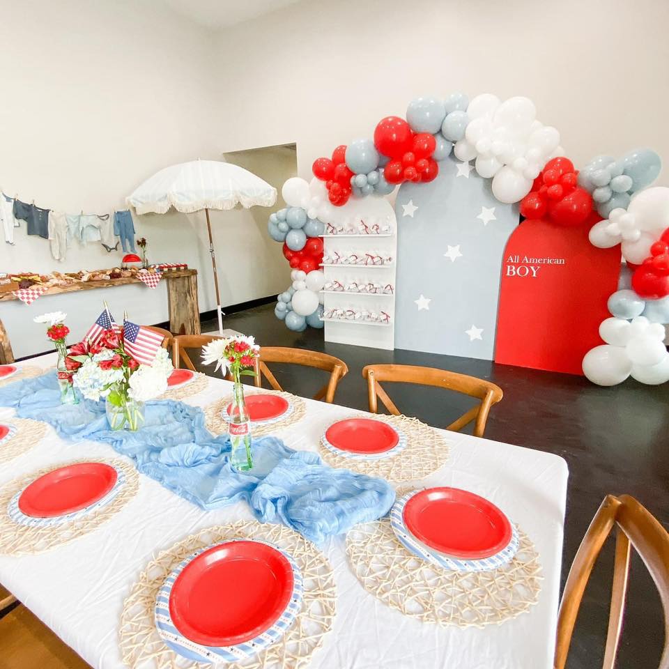 All American Boy theme table setting with red plates and blue table runner under a balloon arch backdrop.