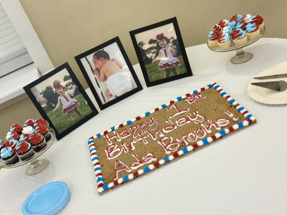 Table display featuring a first birthday cookie cake for Ada Brooks, framed photos, and cupcakes.