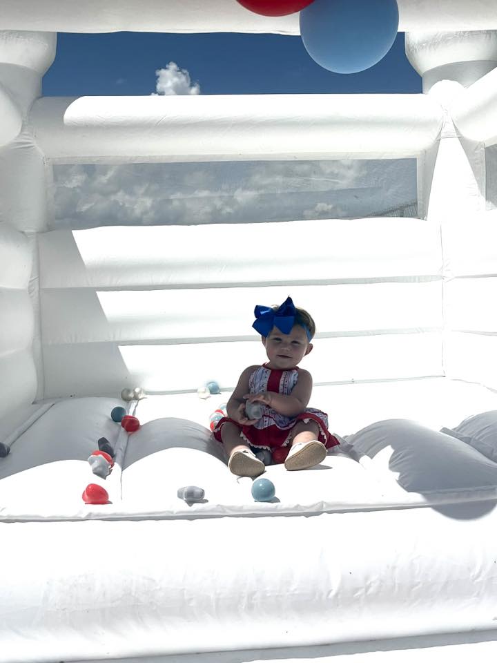 Toddler in a red dress with a blue bow sitting inside a white inflatable bounce house with colorful balls.