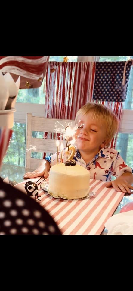 Toddler smiling while looking at a small birthday cake with a lit sparkler candle on a table decorated with a flag
