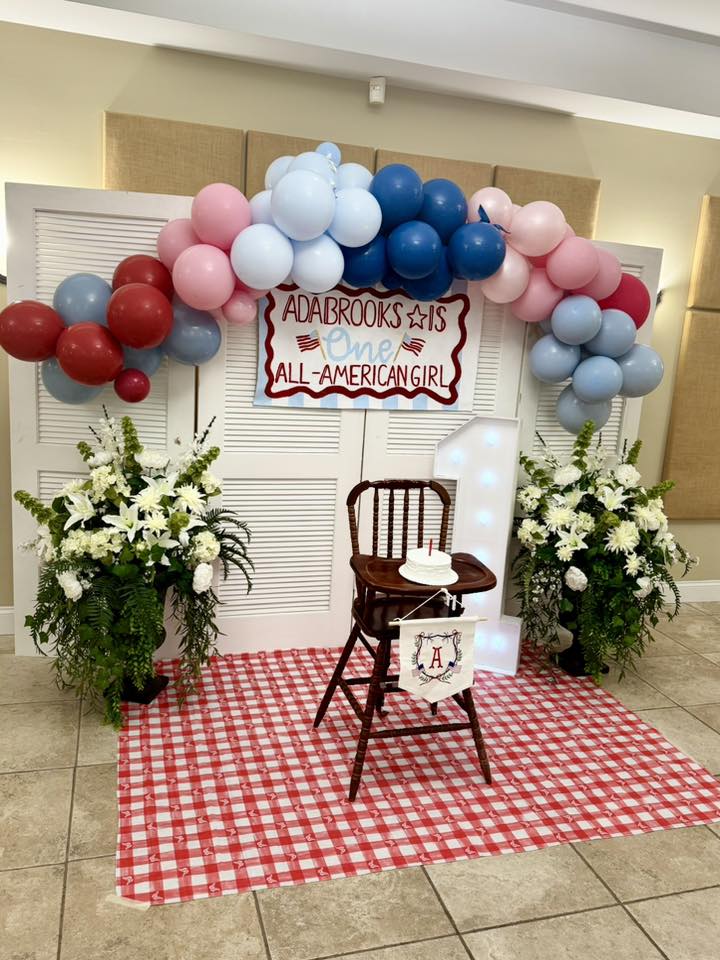 First birthday party setup with red, white, and blue balloon arch and red checkered picnic rug.