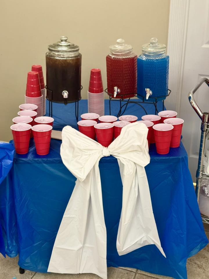 Drink station with three beverage dispensers, stacks of red party cups, and a white bow on a blue tablecloth.