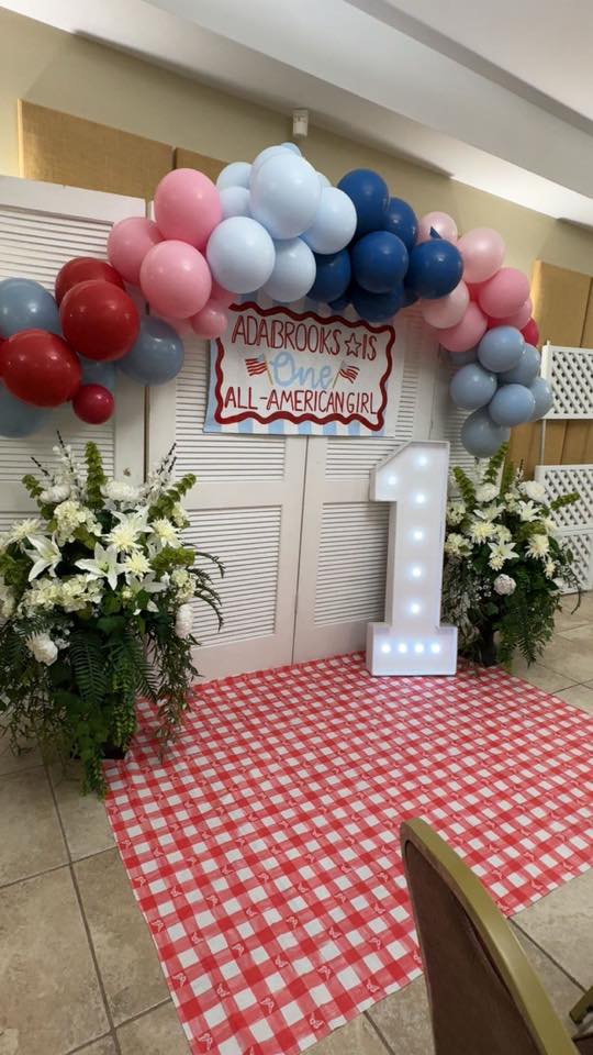 A first birthday party backdrop featuring a red, white, and blue balloon arch, a sign reading Adabrooks is One, and a large light-up number one.
