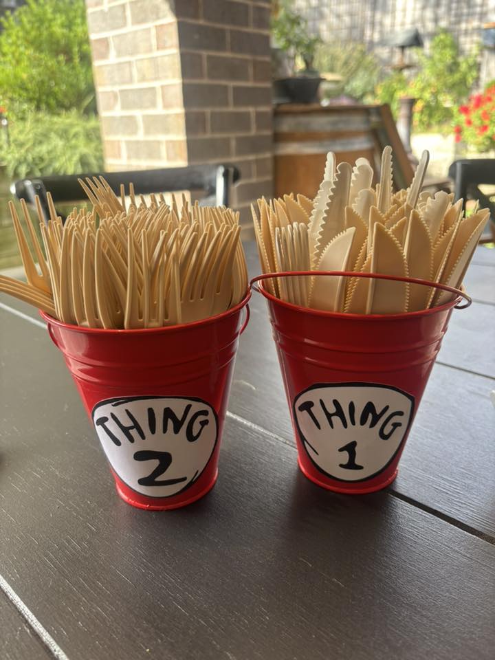 Two red buckets labeled Thing 1 and Thing 2 filled with disposable beige cutlery on a wooden table.