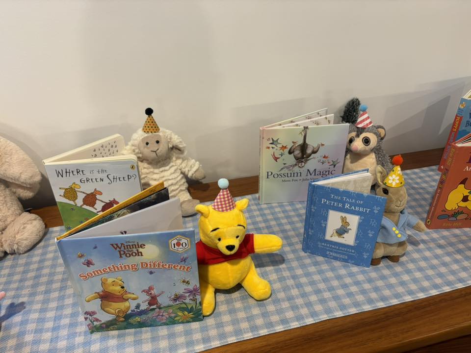 Soft stuffed animals wearing party hats sitting next to classic children's books on a gingham table runner.