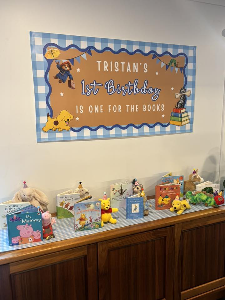 Custom Tristan's 1st Birthday banner displayed above a table filled with children's storybooks and plush characters.