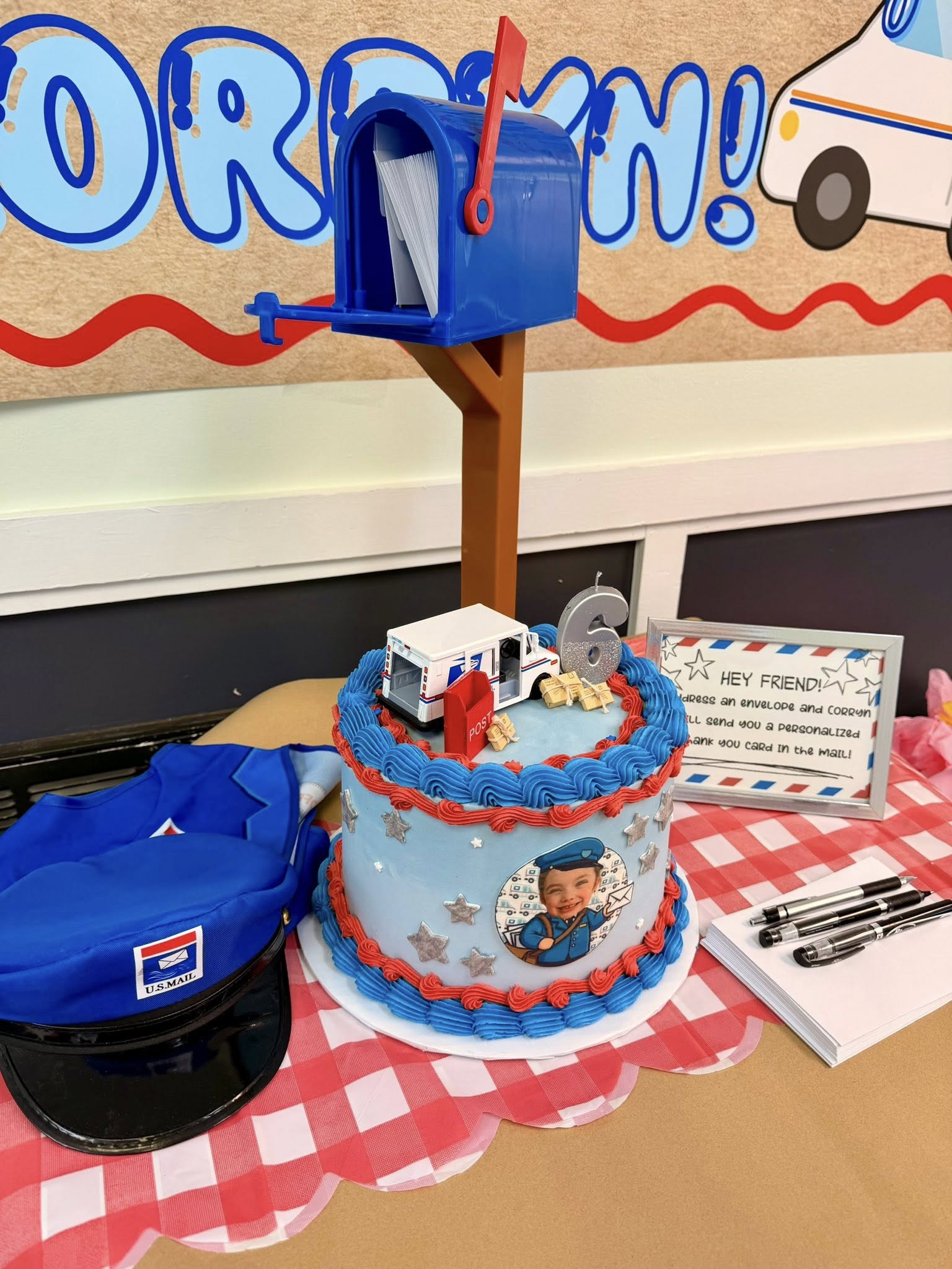 Mail carrier themed birthday cake with a number 6 candle, toy mail truck, and photo topper on a table with a postal hat.