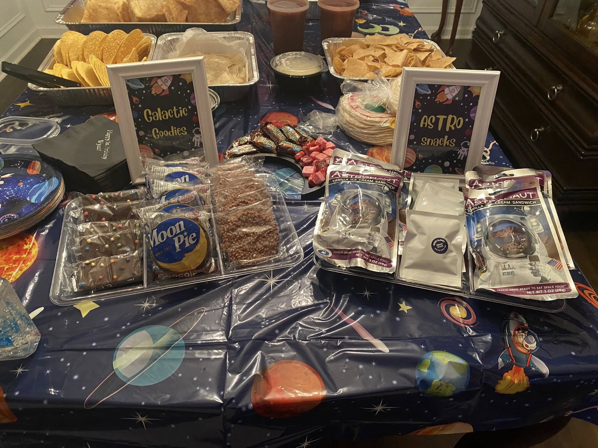 Space-themed party food table with Moon Pies, astronaut ice cream, and signs for Galactic Goodies and Astro Snacks.