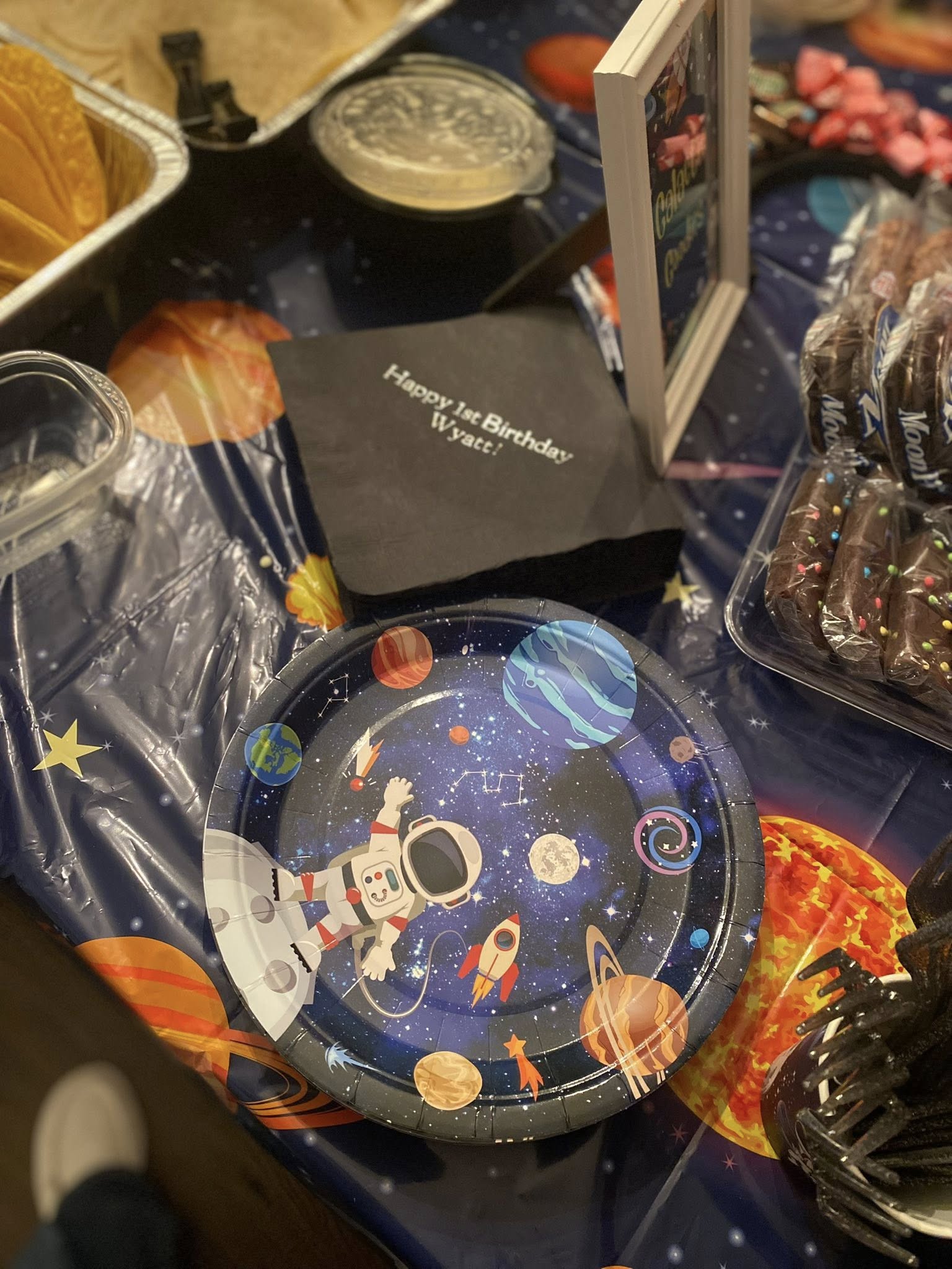 Space-themed party paper plate next to napkin that says Happy 1st Birthday Wyatt on a galaxy tablecloth.