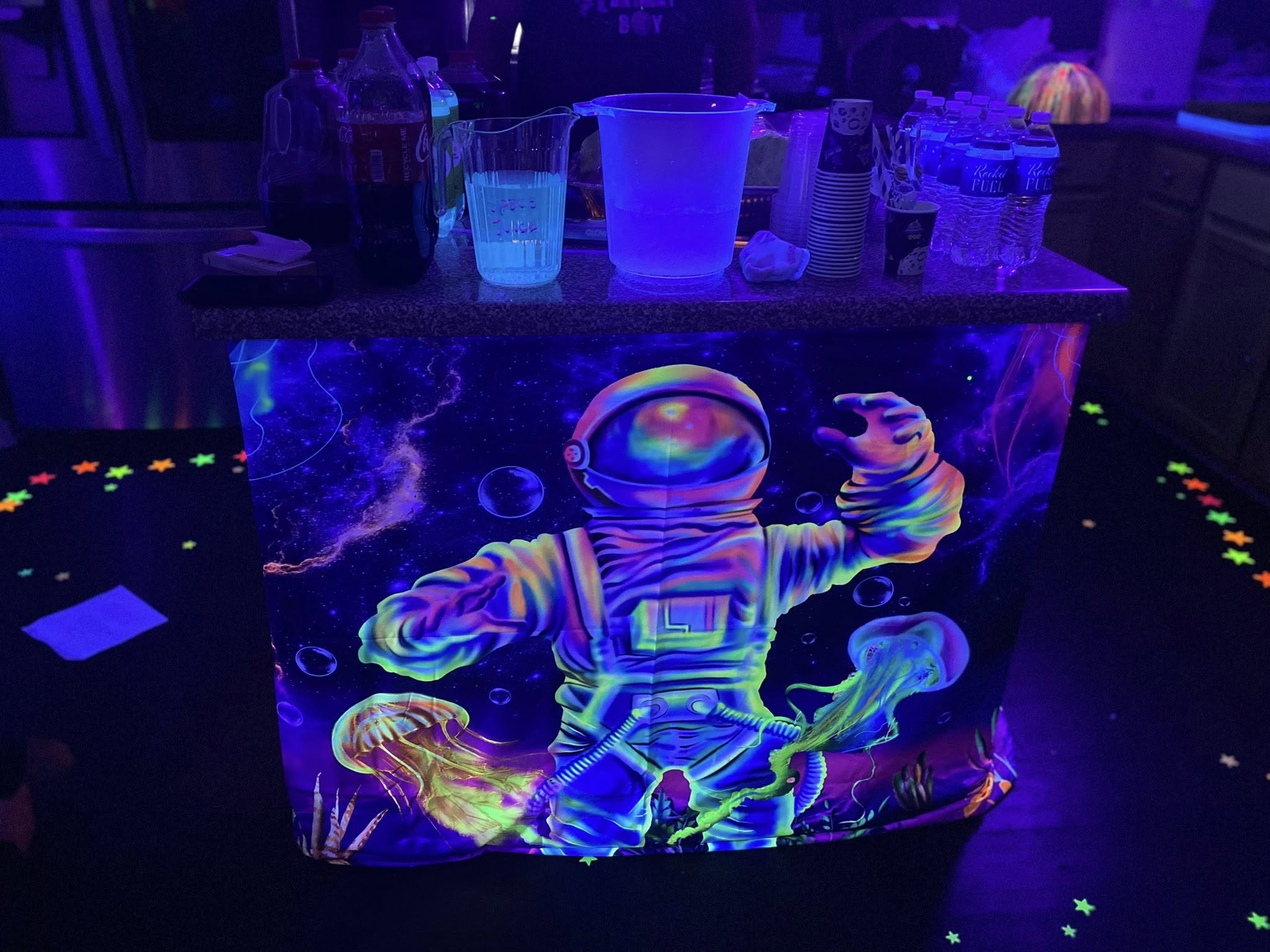 Table covered in a glowing neon astronaut and jellyfish tablecloth at a party with drink supplies on top.