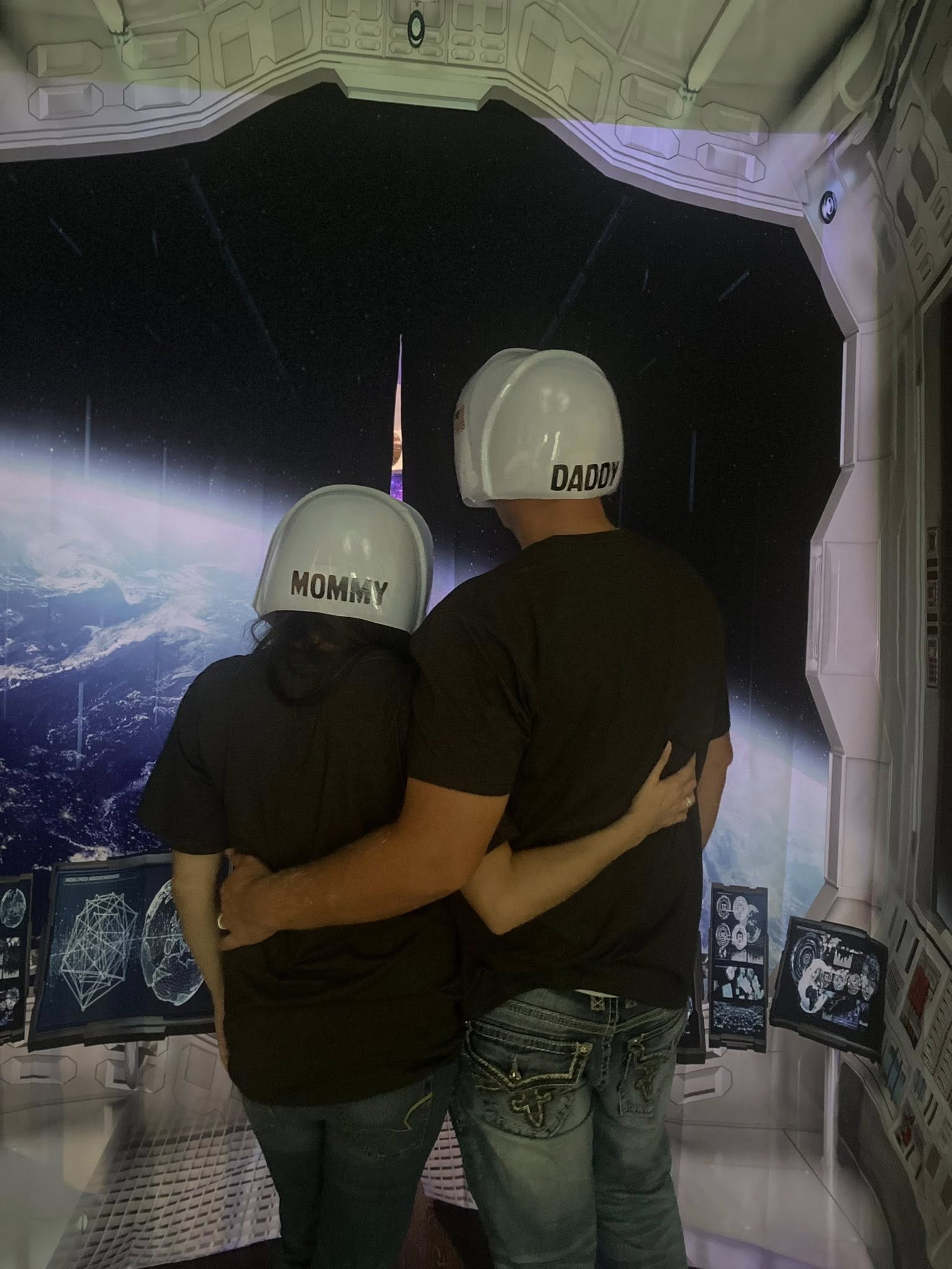 Couple wearing astronaut helmets labeled Mommy and Daddy looking out at a space scene on a digital display