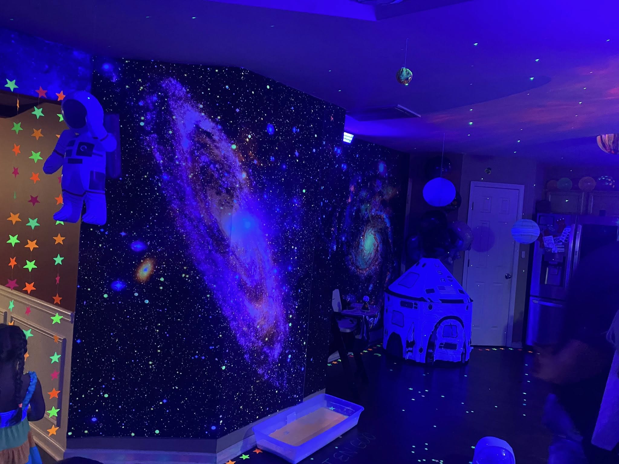 Room decorated with blacklight space-themed wall murals, hanging astronaut cutout, and a NASA play tent.