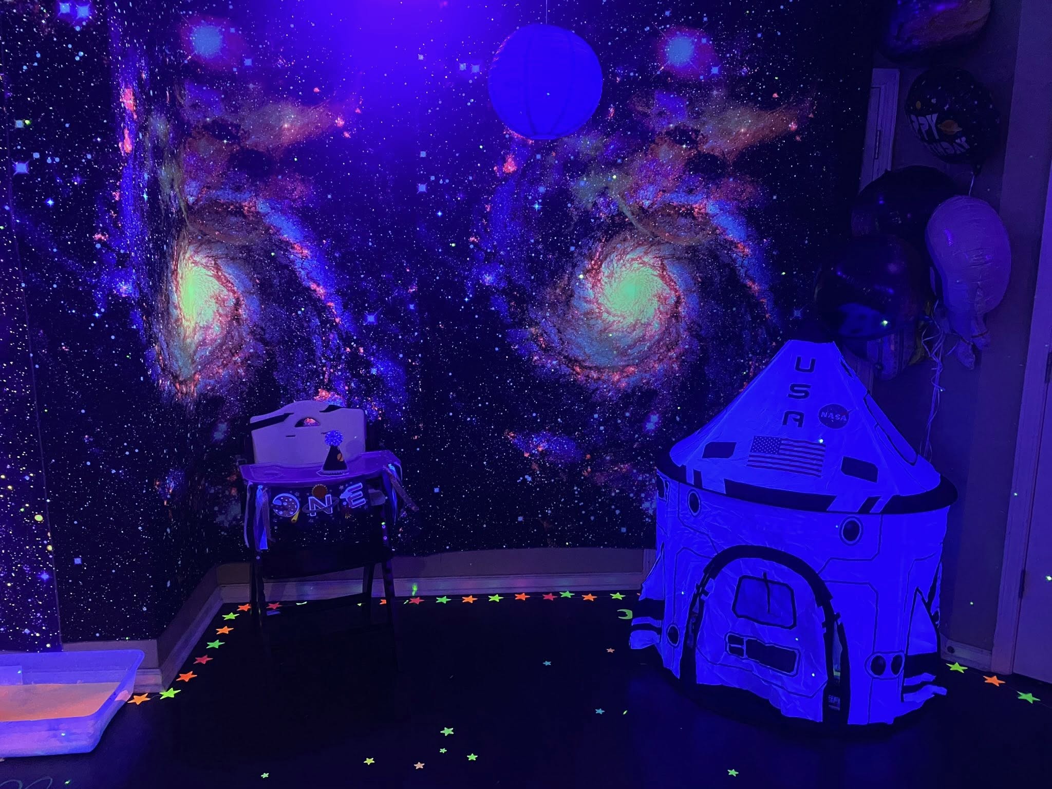 Space-themed first birthday party setup with galaxy backdrop, toddler high chair, and NASA play tent.