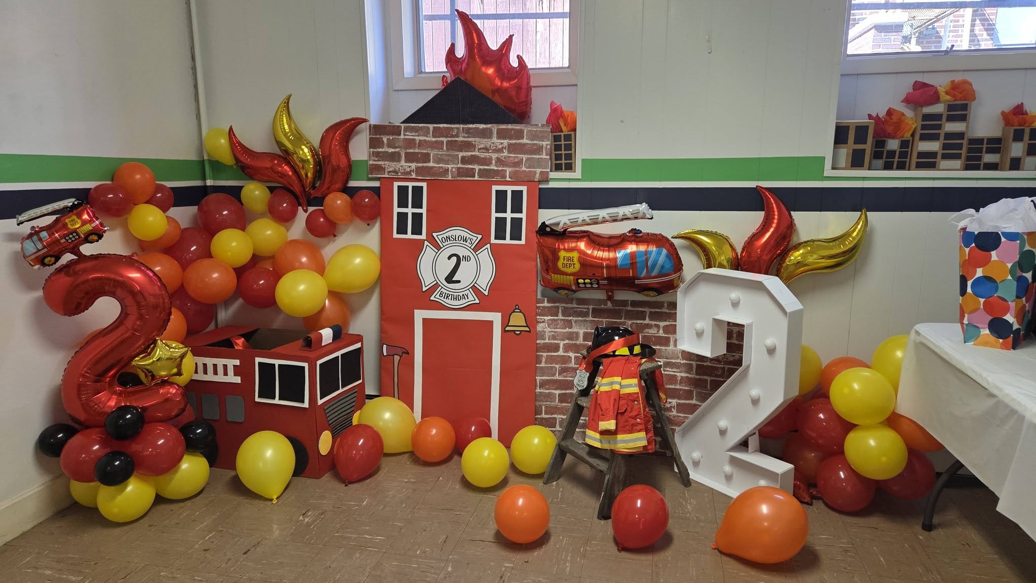 Firefighter themed second birthday party decorations featuring a fire station backdrop, balloons, and a number two light.