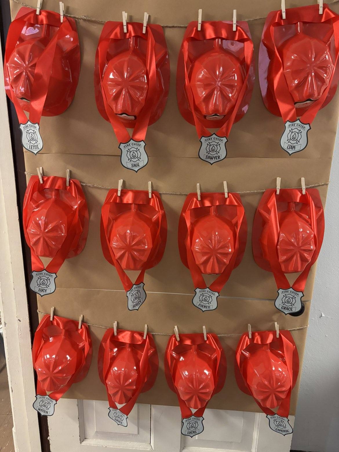 Personalized plastic red firefighter hats with name badges hanging on a wall for kids party favors