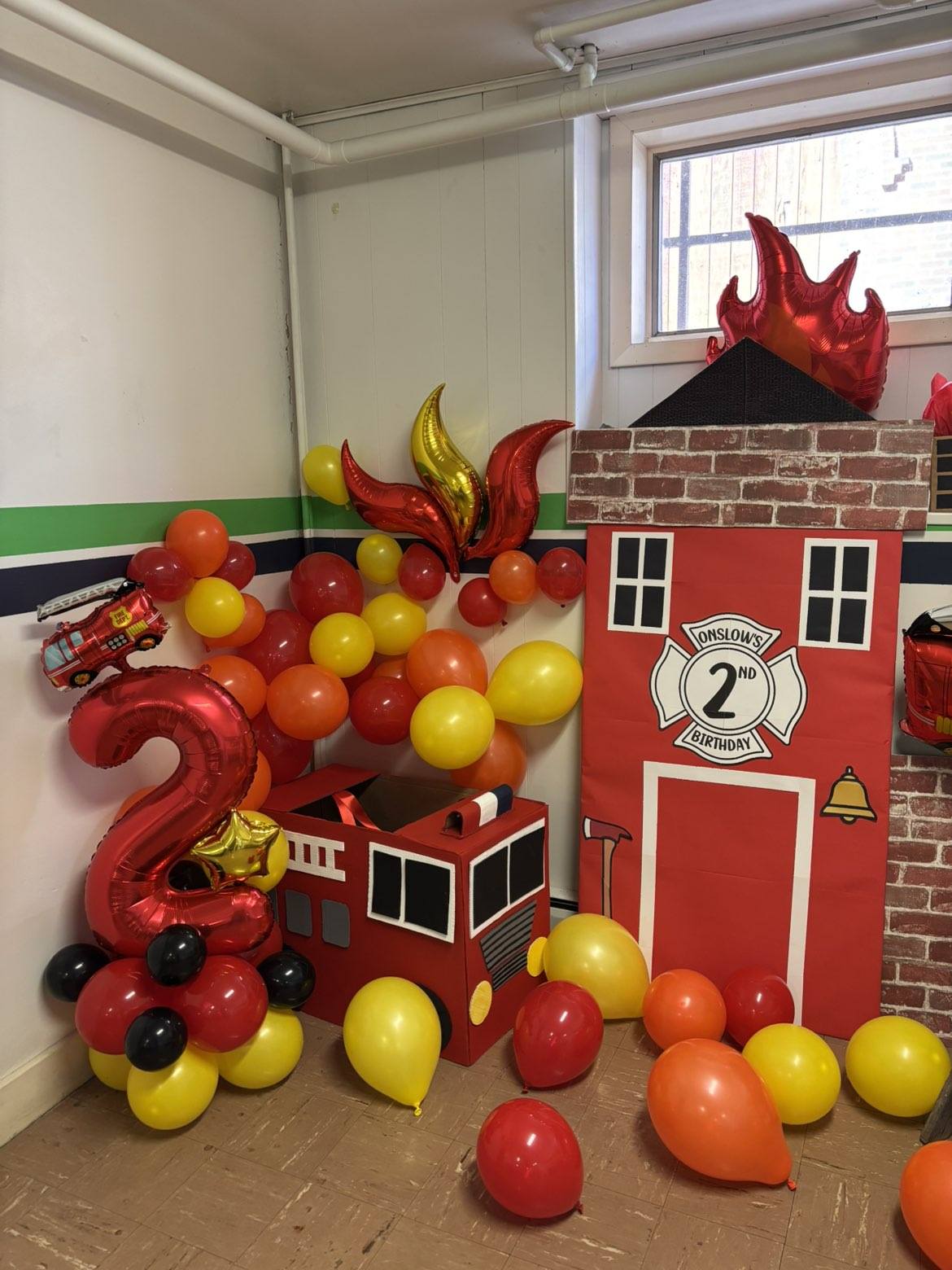 Firefighter themed birthday party decorations with red and yellow balloons, a cardboard fire station, and a number 2 balloon.