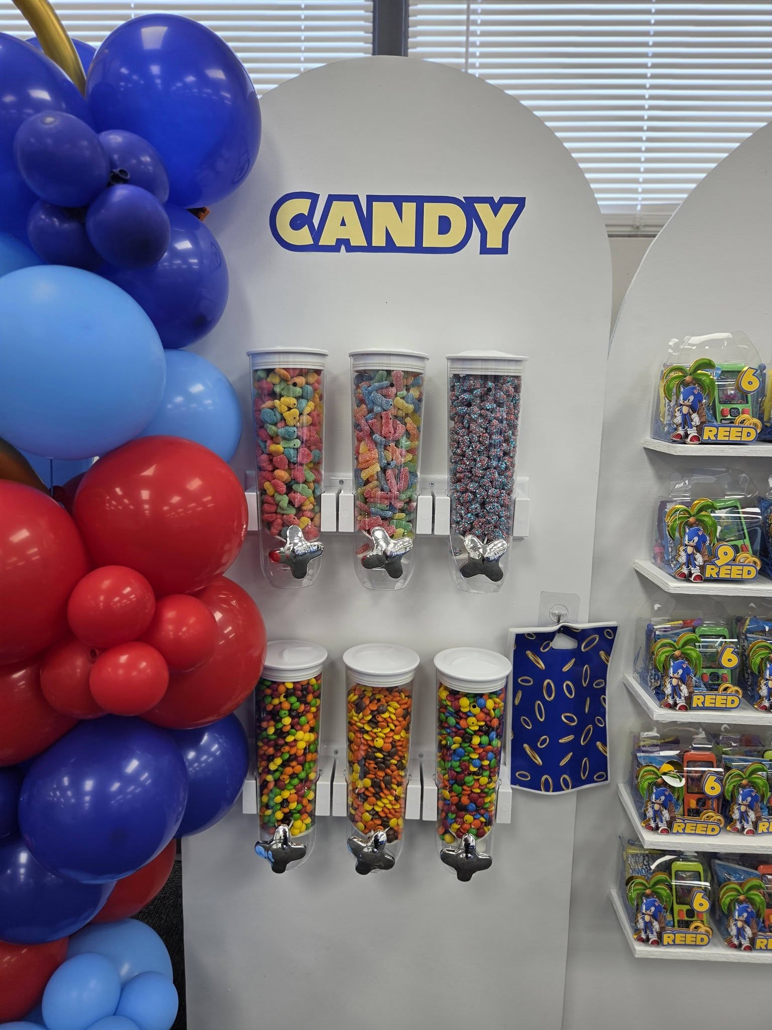 Candy dispenser station with rows of colorful gummies and chocolates decorated with blue and red balloons.