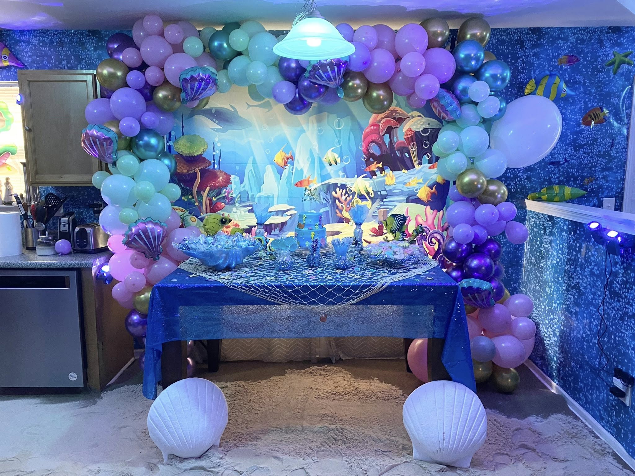 Table set up for under the sea mermaid themed birthday party with blue balloon arch and seashell decorations.