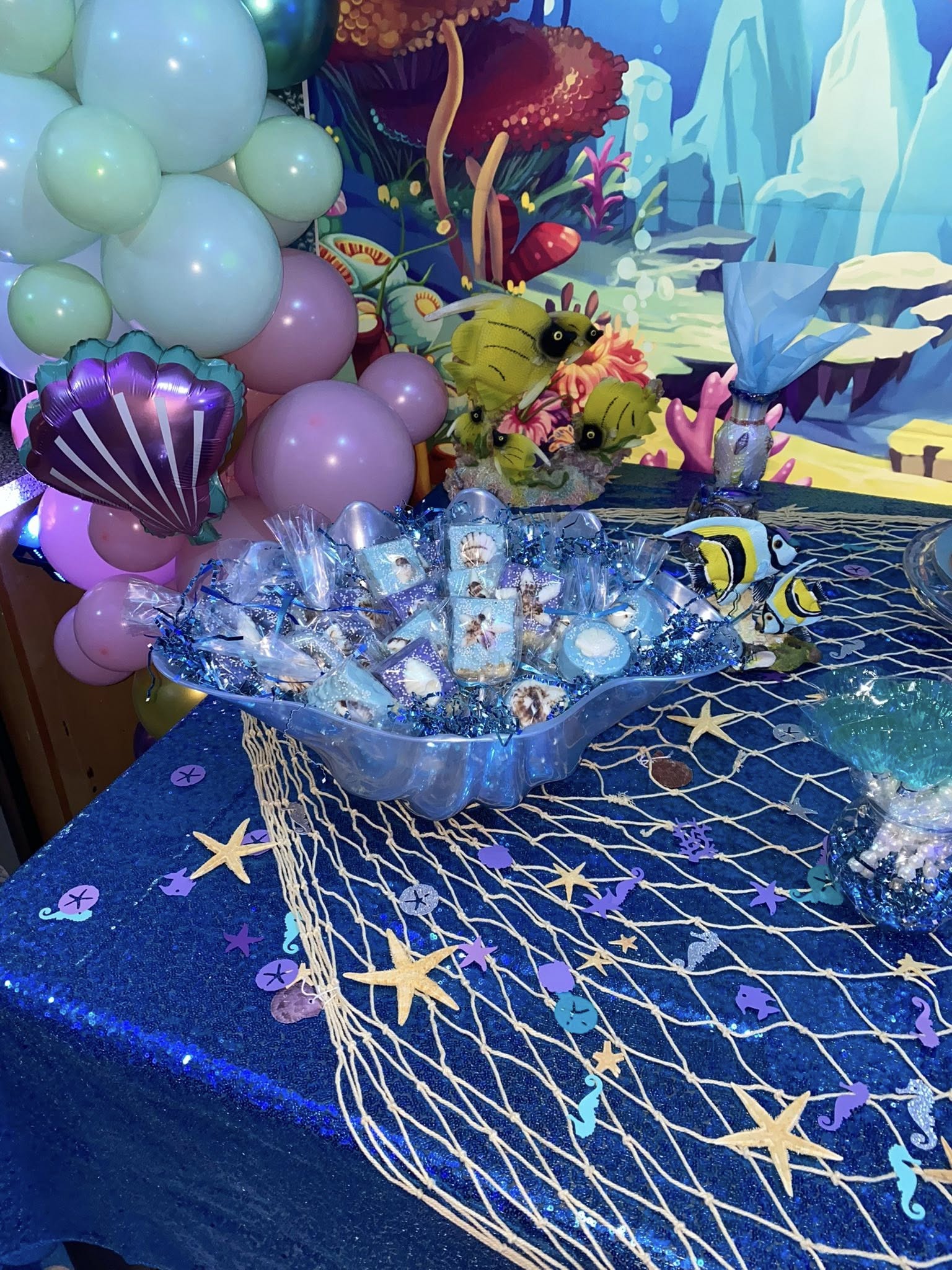 Table display with scattered individually wrapped ocean-themed party favors in a shell-shaped bowl, atop a blue sequined cloth with a fishnet runner.