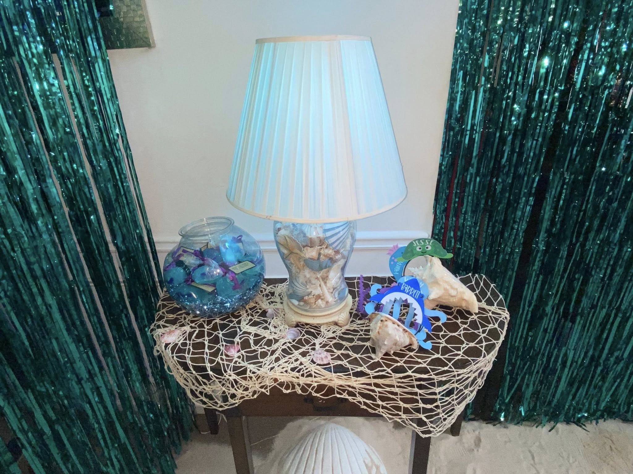 Nautical themed tabletop lamp with seashell base, fish bowl, and net decoration against teal fringe curtains.