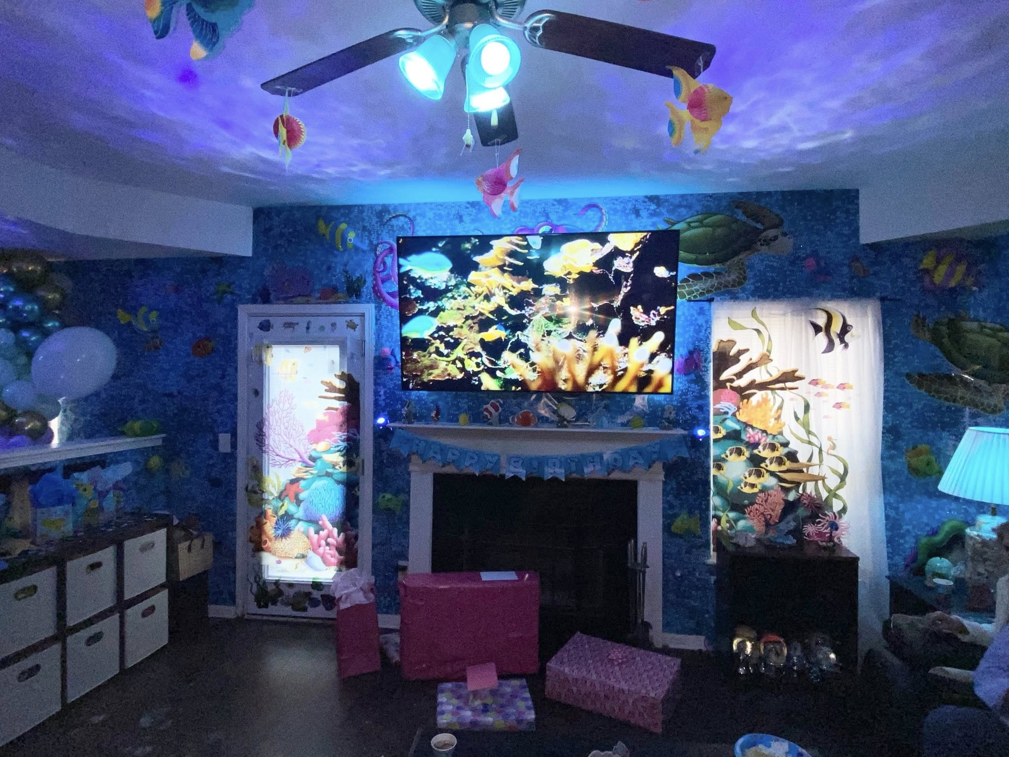 Living room decorated for a birthday with an ocean-themed wall mural, blue ambient lighting, and gift boxes.