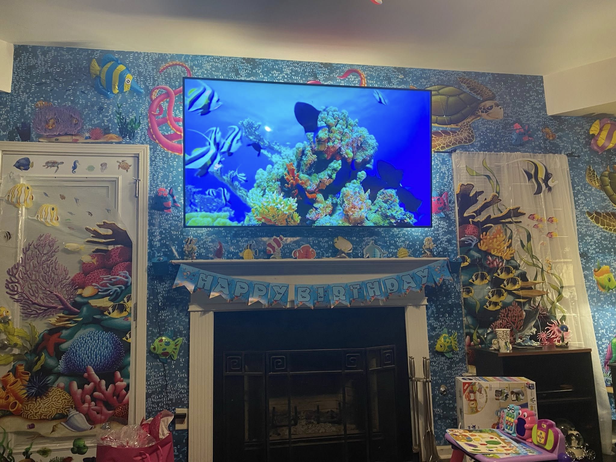 Living room fireplace wall with ocean-themed wallpaper, a flat-screen TV displaying a coral reef, and a Happy Birthday banner.