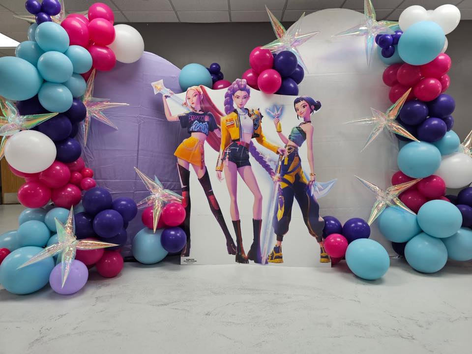 Cardboard cutout of three K-pop themed characters surrounded by a colorful balloon arch and holographic star accents