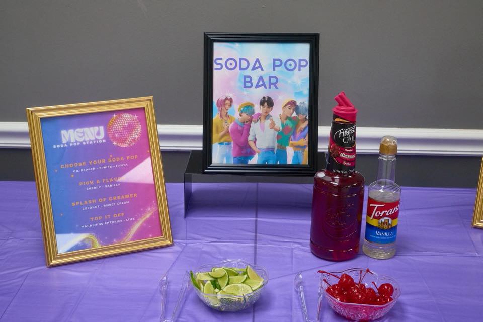 Soda pop bar station with menu sign and assorted drink garnishes including lime slices and maraschino cherries.
