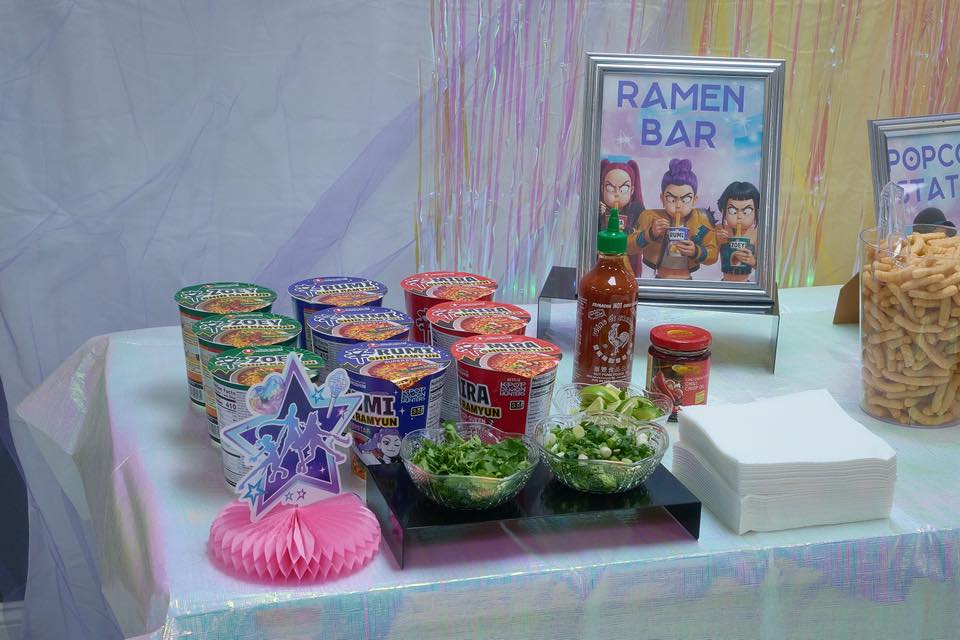 Table set up as a ramen bar with variety of instant ramen cups, sriracha, fresh toppings, and themed signage.