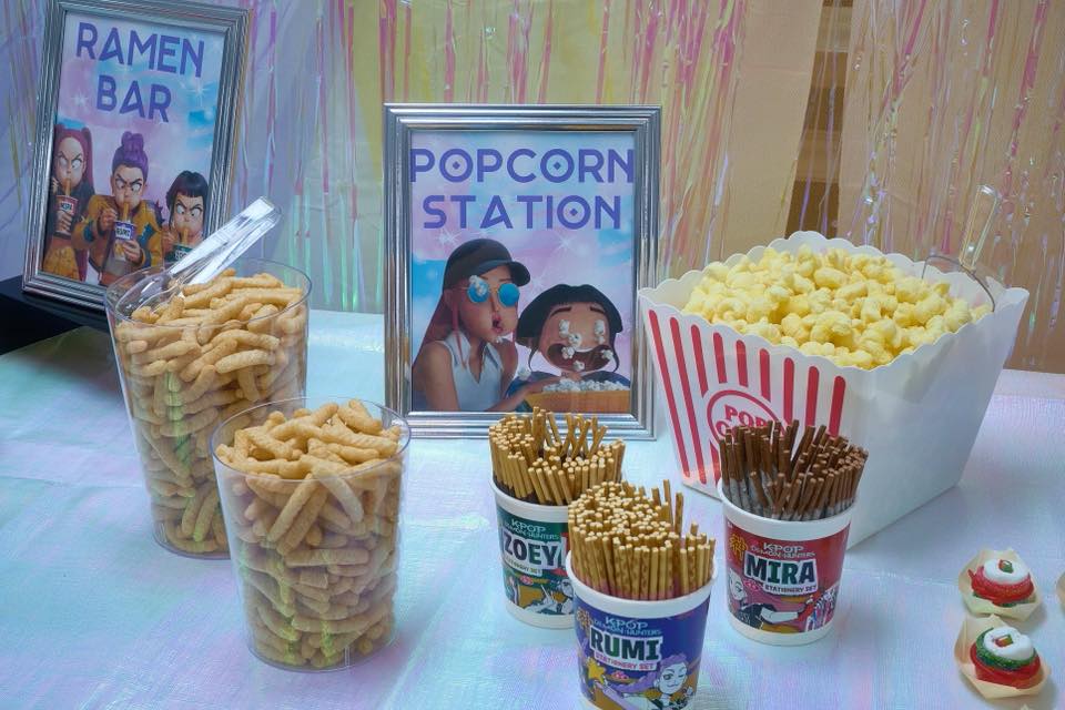 Table set up with snacks including Ramen Bar and Popcorn Station signs, popcorn in a red and white striped container, and various pocky stick cups featuring K-Pop character themes.